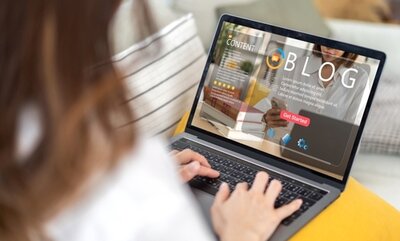 Picture of a woman sitting on a couch with her laptop on her lap. Laptop is pulled up on a page that's says blog.